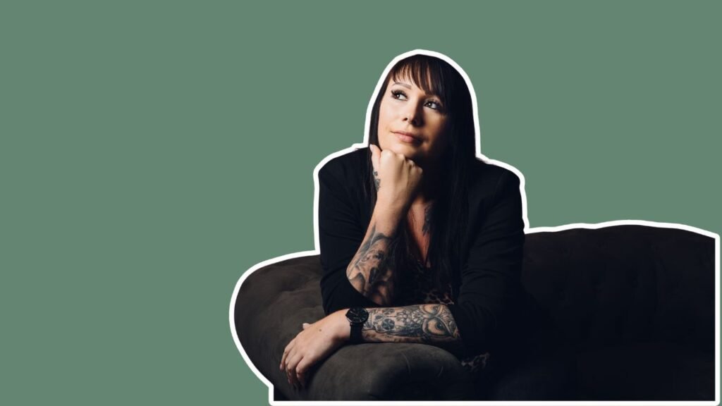 Person mit tätowierten Armen sitzt nachdenklich auf dunklem Sofa vor grünem Hintergrund.